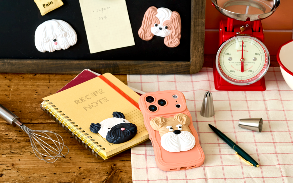 image set in on a kitchen countertop shows two of the Patisstickers mounted to a black board, a fluffy white one and tan and white long eared one. Atop a yellow notebook, beside a metal whisk and peach cell phone sits the tan and white short nosed dog, a black pen and red kitchen scale