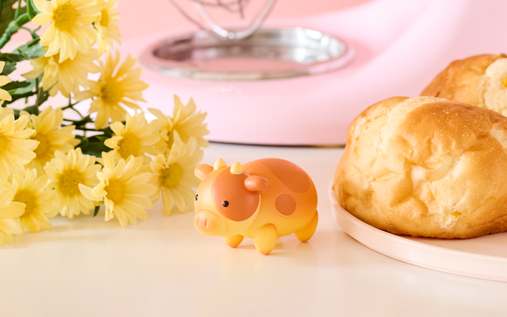 Small yellow animal figurine next to a plate of bread and yellow flowers on a pink surface.