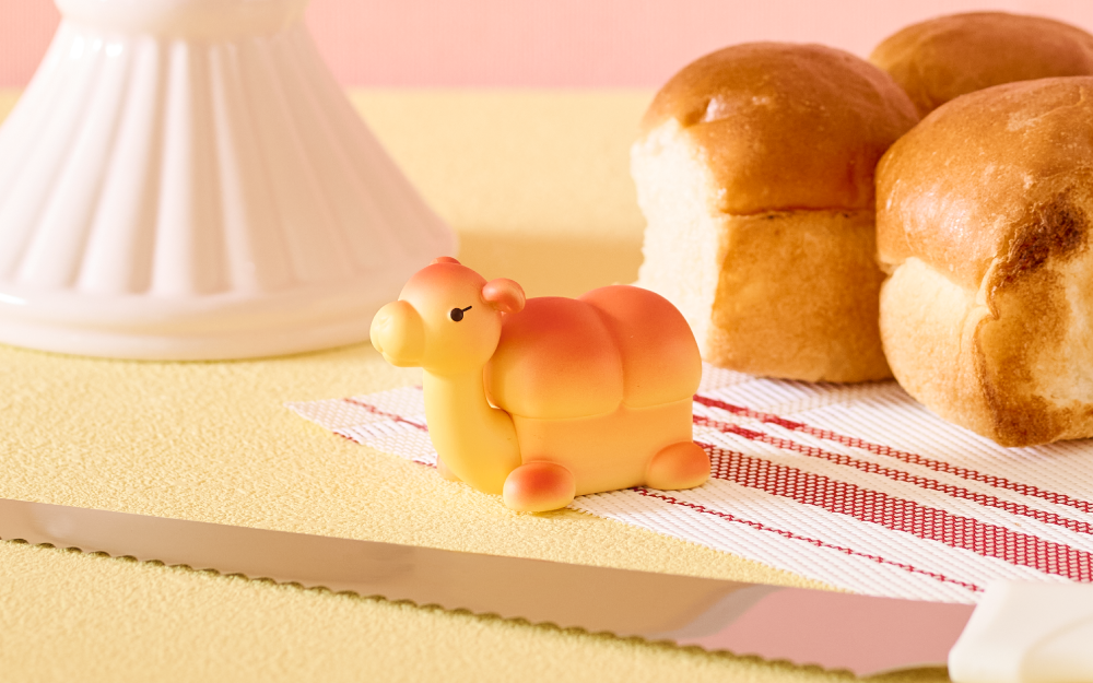 Small yellow camel-shaped minifigure on a pink surface with bread loaves in the background