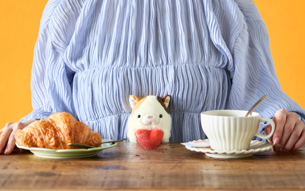 Set against a rich orange background, image shows figure wearing a blue striped shirt sat at a wooden table. Atop the table is a croissant, and coffee cup and saucer. In the center of the image, poised in the figure's lap is the Calico variant of the Posture Pal Bag Charm holding a plush red heart. 