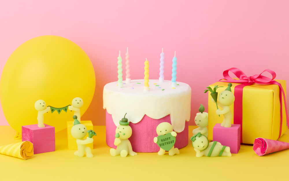 Image set on pink background with yellow balloon, white frosted cake with candles and yellow gift. Image shows the new product line including SMISKIs holding a banner, SMISKI in party hat holding a bouquet, SMISKI with a cherry on head eating frosting, SMISKI holding heart with "Happy Birthday", SMISKI with a candle flame atop its head, SMISKI holding a confetti popper with birthday hat, and SMISKI lying down wrapped in ribbon.