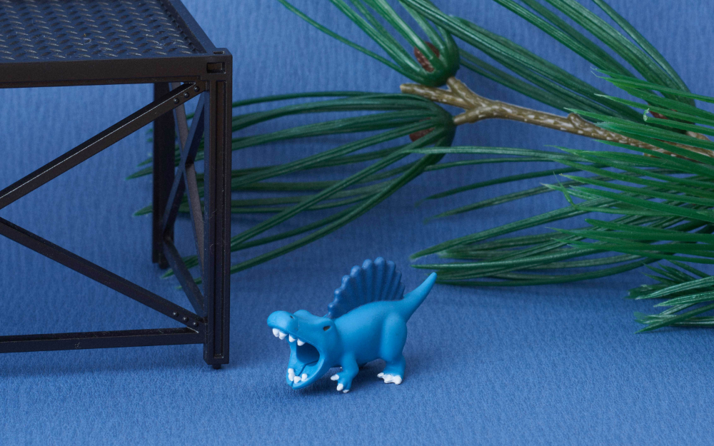 product lifestyle image of cable bite dino spinosaurus with a fake pine tree and scaffolding prop against a blue background