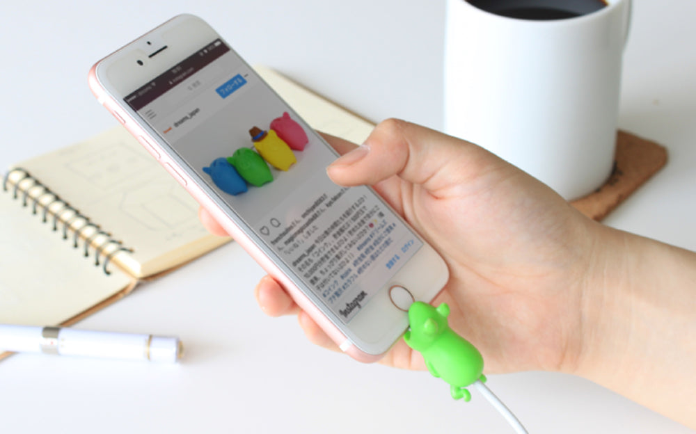 Image shows a hand holding a cell phone with the Cable Bite Volume 1- Frog variant seen on a charging cable attached to the phone. Behind the phone, an open notebook and cup of coffee can be seen