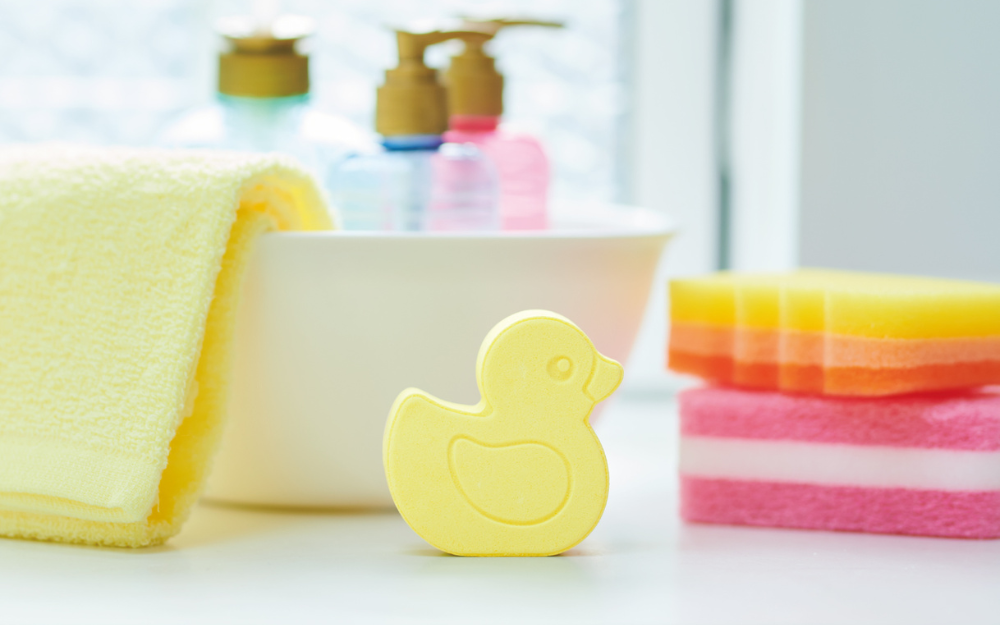 lifestyle image of rainbomb mini yellow duck on a white sink background with bath sponge, soap bottle, and towel props