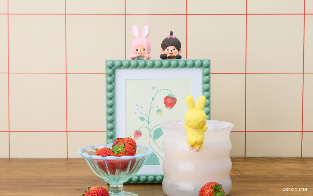 Two Monchhichi Hippers; pink bunny and girl figure are set atop a teal framed image of a strawberry, against a red and white background. Set in front is a bowl of strawberries, as well as the yellow bunny HIPPER facing away, set on a white mug. 
