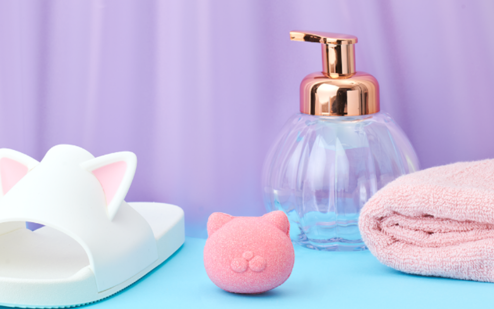 Set on a purple background, a Nekomote Rose bath bomb sits on a blue counter top beside a white cat slide, clear pump dispenser and pink folded towel