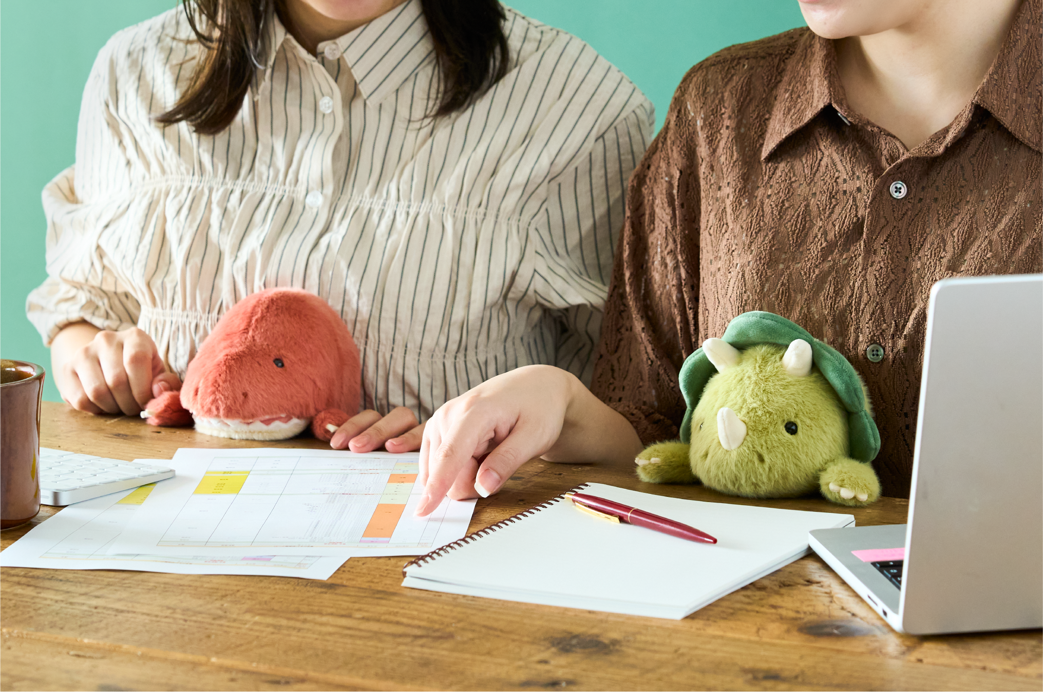 Set on a teal background. Two figures, one in white and gray striped shirt, the other in a brown pattern button down shirt sit side by side at a desk looking over a notebook and papers. A laptop, keyboard and mug are set on the desk with them. On left, light red and white Tyrannosaurus Rex Posture Pal supports one figure, and the other holds the light and dark green Triceratops Posture Pal