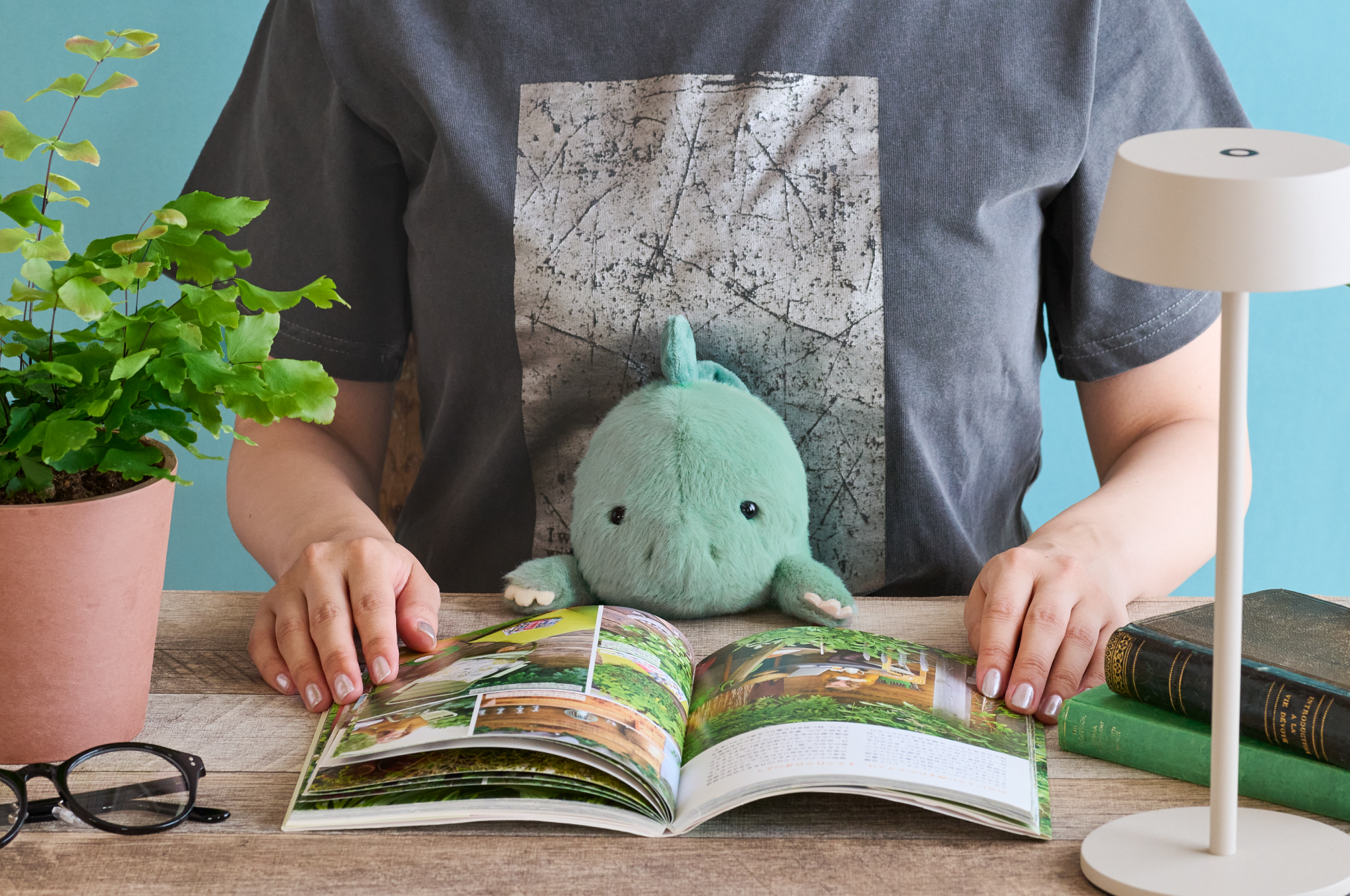 Image set on light blue background. Figure sits in grey and white shirt with light and dark teal Stegosaurus Posture Pal with open gardening book in front of them. Beside figure on table sit potted plant, pair of glasses, small table lamp and stack of books. 