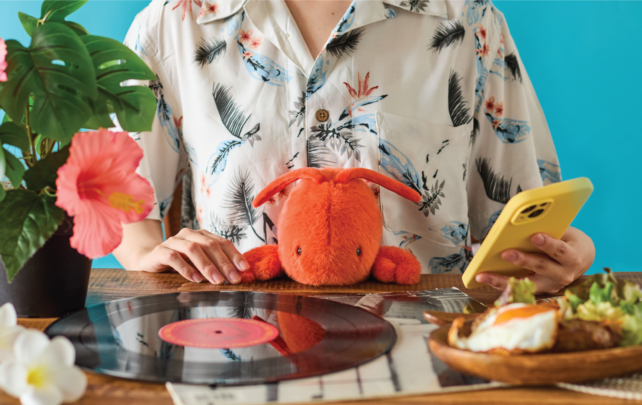 Image shows the upper body of a person in a floral button down shirt, holding a yellow cell phone, a red/orange Lobster Posture Pal sits in their lap. The wooden table is set with a vinyl record, flowers, potted pink flowering plant, and plate of food. Image is set on a bright blue background.