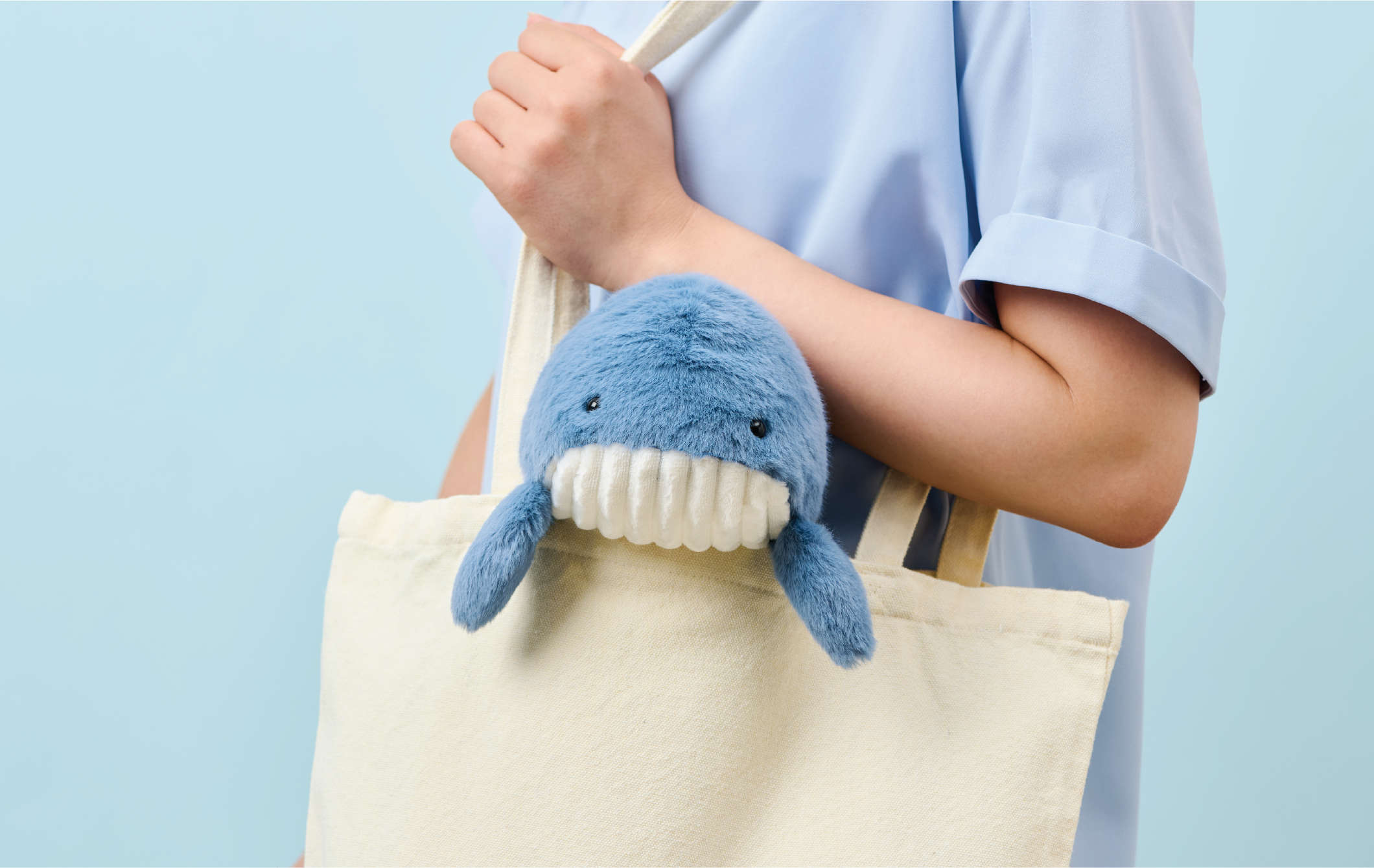 Image shows the upper body and forearm of a person in a blue shirt holding a white tote bag with blue Posture Pal whale inside set against a light blue background
