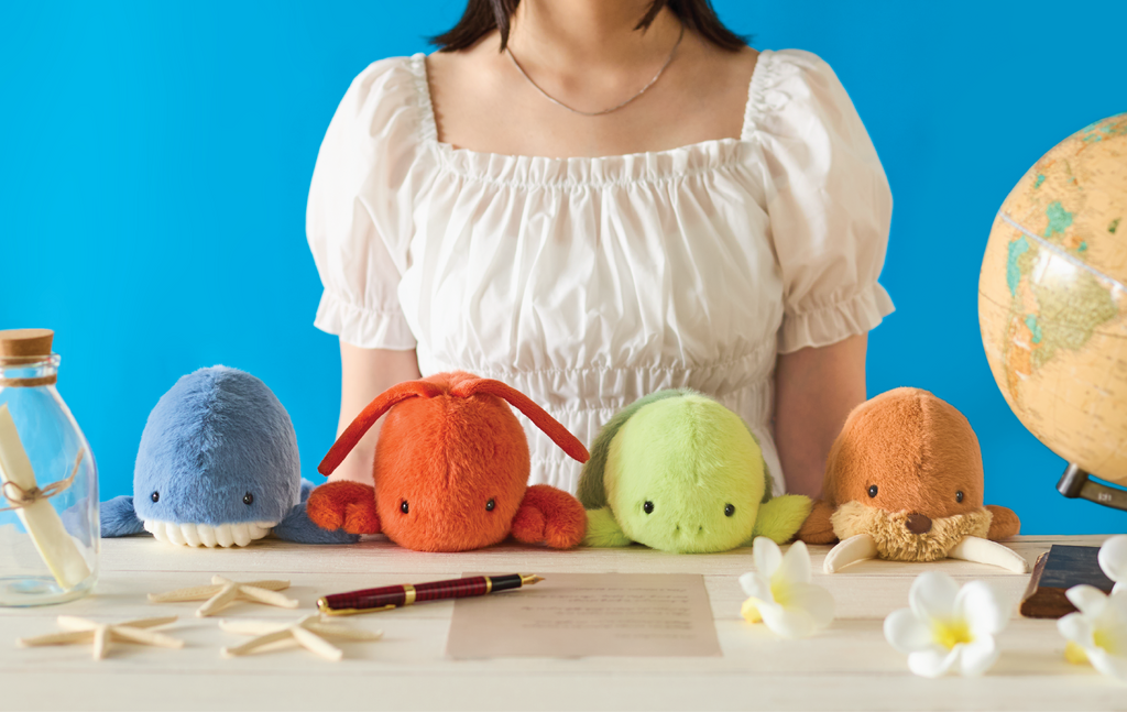 Image shows the upper body of a person in a white shirt sitting with colorful Posture Pals- left to right: blue whale, red/orange lobster, green turtle, brown walrus and a globe, flowers, starfish, a pen and message in a bottle sit on a table against a blue background