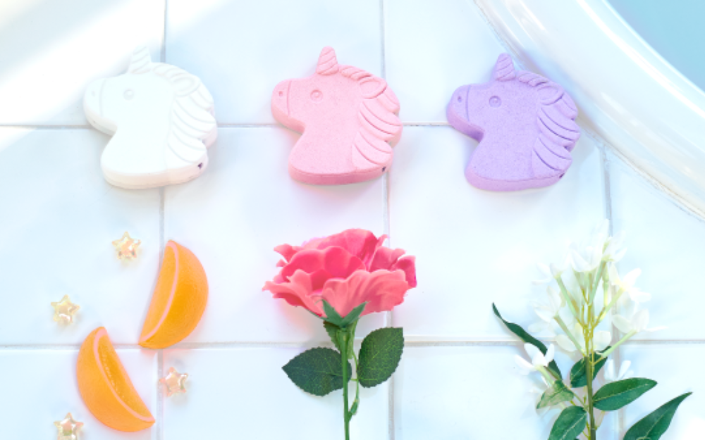 lifestyle image of rainbomb unicorn, sweet rose, and jasmine (left to right) on a white tile backdrop above orange slice, rose, and jasmine props