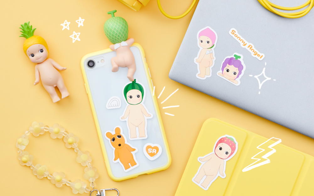 product lifestyle image of the sonny angel sticker pack featuring die cut stickers from the fruit pack placed on top of electronic items such as a cell phone case, laptop front, and tablet case. the overall theme of the image is YELLOW and there is one sonny angel pineapple mini figure in the upper lefthand corner and one melon hipper attached to the back of the phone case.
