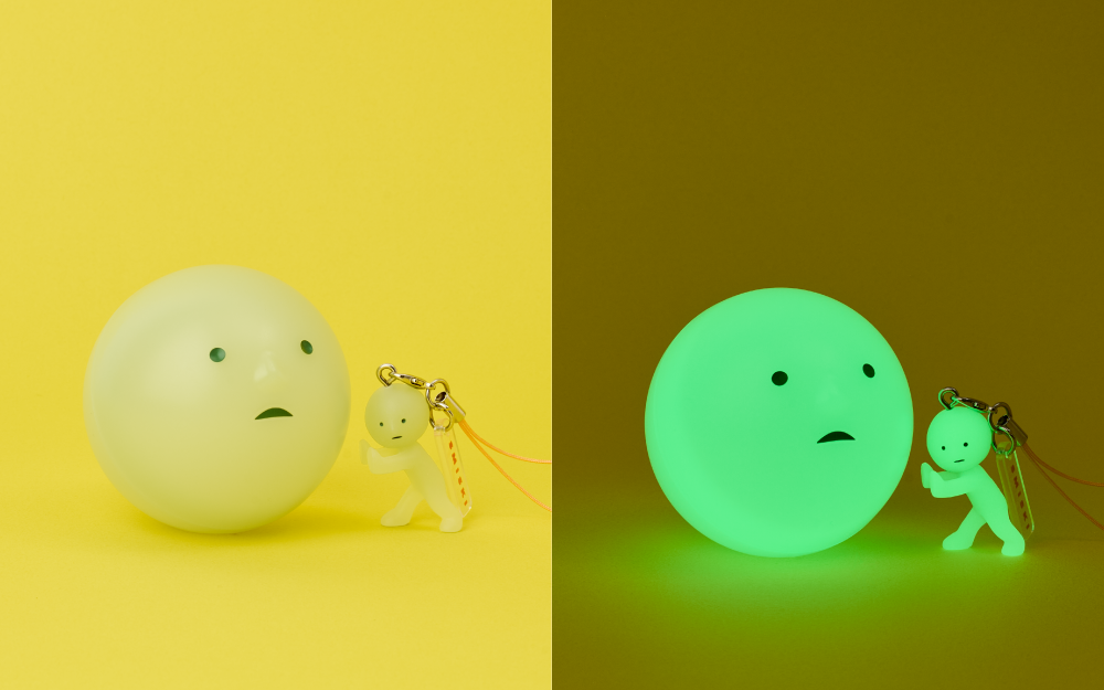 Split image shows the SMISKI Accessory capsule product in daylight and while glowing. Left side: set against a yellow background, the SMISKI capsule is set beside the SMISKI Pushing charm variant. Right: The same capsule and figure stand in a low light setting, showing the glow in the dark feature of both the capsule and charm