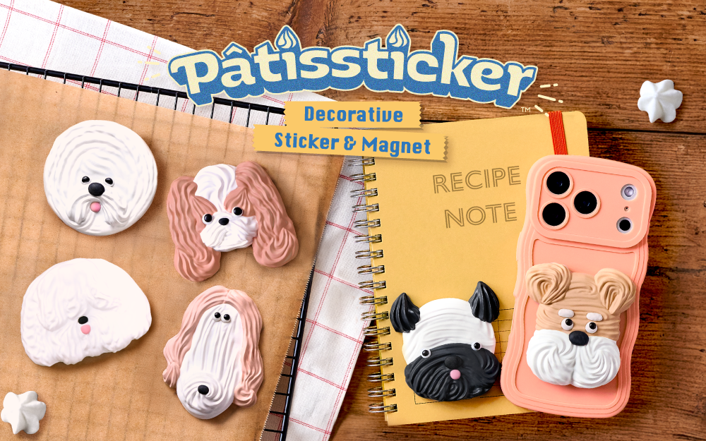 new product image set against wooden background introduces Patissticker Decoration Sticker and Magnet. Atop a cookie sheet from left to right, a white round dog, tan and white long eared dog, fluffy white dog, long faced tan and white dog, black and white short nosed dog on a notebook and tan and white short hair dog on the back of a peach cell phone case