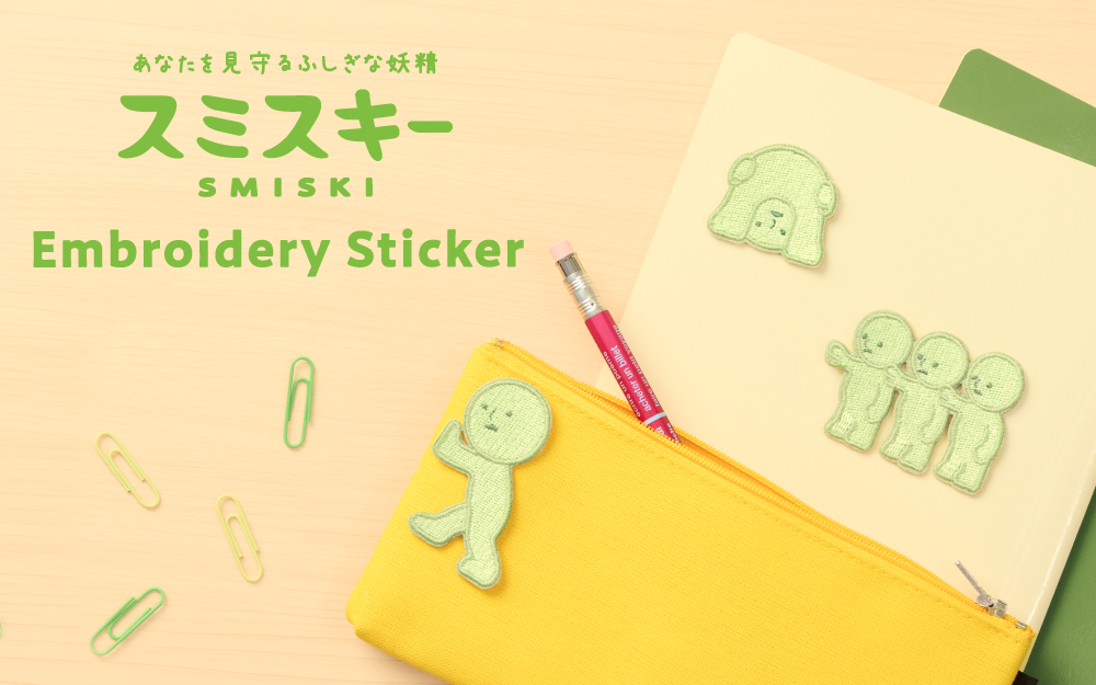 SMISKI Embroidery Sticker text set on wooden background with green paperclips, yellow pencil pouch with Pushing SMISKI sticker on left side. Red pencil poking out of pouch, with tan folder and Peeking 2, and Little variants atop a green folder. 