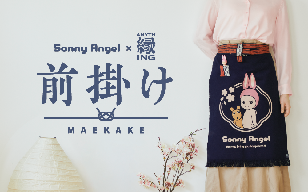 White background with navy blue text "Sonny Angel and Anything" collaboration "Maekake" apron. Below text is a paper lantern, cherry blossom plant, and person. Person is wearing a pink shirt, tan skirt and navy blue apron. Apron has red and white striped ties, navy fringe, wrapped around the waist of the figure. Red and white striped pocket holds Rabbit Sonny Angel minifigure. Apron design: Rabbit Sonny Angel beside Robby encircled with cherry blossoms, and text: "Sonny Angel" "He may bring you happiness?!"