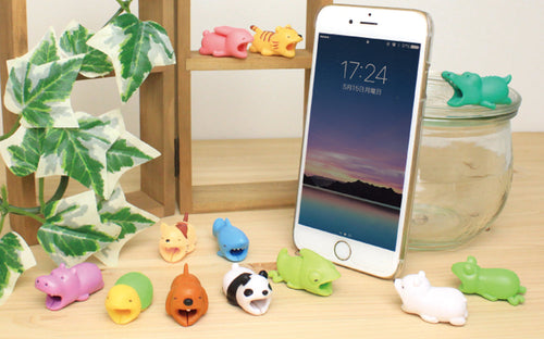 Image shows all product variants for Cable Bite Vol. 1 Cable Accessory. On top of a wooden frame shelf, Rabbit and Tiger. On a wooden tabletop beside green ivy, Hippopotamus, Turtle, Dog, Panda and Chameleon; in the row behind: Cat and Shark. To the right of an white iPhone, Polar Bear, Frog and above on a glass dish, Crocodile